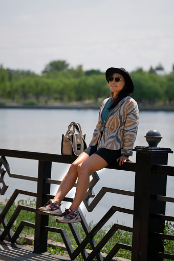 Portrait of a young Asian (Kazakh) woman in sunglasses and a hat. Summer urban portrait of a happy girl of beautiful oriental appearance. The girl is standing on the embankment of the river.