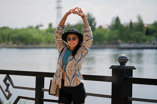 Portrait of a young Asian (Kazakh) woman in sunglasses and a hat. Summer urban portrait of a happy girl of beautiful oriental appearance. The girl is standing on the embankment of the river.