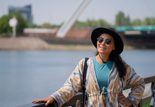 Portrait of a young Asian (Kazakh) woman in sunglasses and a hat. Summer urban portrait of a happy girl of beautiful oriental appearance. The girl is standing on the embankment of the river.