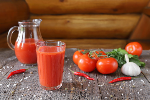 A tasty tomato juice in a glassware and fresh tomatoes with vegetables, herb and spicy are on a wooden background.