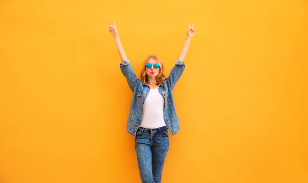 Trendy happy cheerful caucasian young woman having fun raising her hands up wearing denim jacket on yellow background stock photo