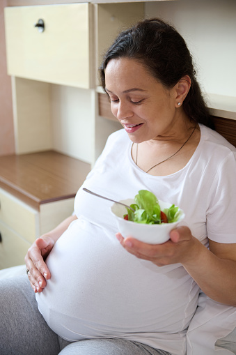 Happy pregnant woman in third trimester of her pregnancy, sitting on the bed, caressing her belly, smiling and eating healthy vegetable salad, expressing positive emotions from her maternity lifestyle
