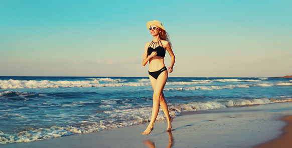 Summer vacation, happy smiling woman walking running in bikini swimsuit and straw hat on the beach on sea coast with waves background on sunny day