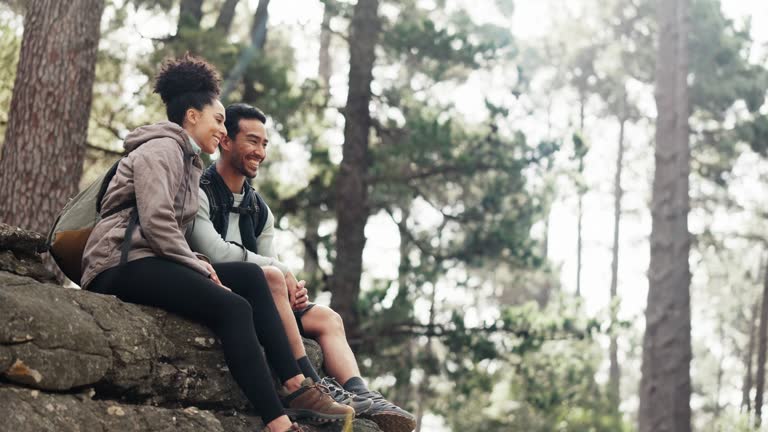 Nature, hiking and a couple relax on adventure trail in forest and sitting on a rock. Health, happy man and woman pointing at natural landscape while relaxing in woods with trees, fitness and freedom