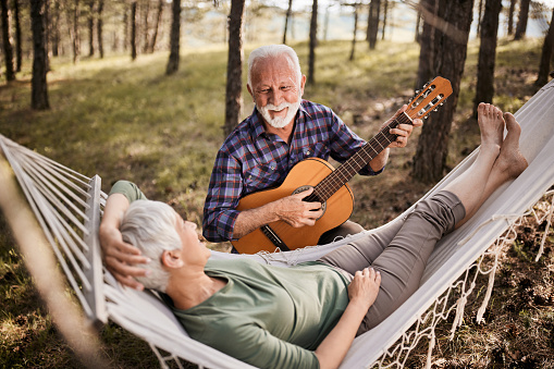 Happy senior man playing a guitar to his relaxed wife in hammock at the backyard.