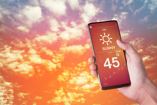 Hand holding smartphone with weather hot sunny day on screen and sky sunlight background.