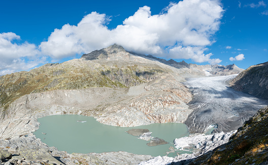 View of the glacier and its glacial lake and the nearby mountains under a blue sky and some clouds. The glacier is located near the famous Furka Pass and the Hotel Belvedere