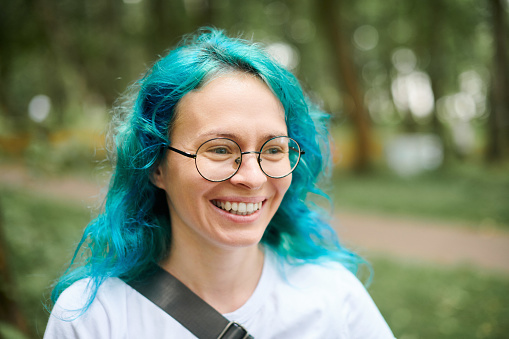 Young adult woman portrait in round glasses with turquoise dyed hair on green forest background, attractive female artist with bright appearance. Charming woman with marine style turquoise dyed hair