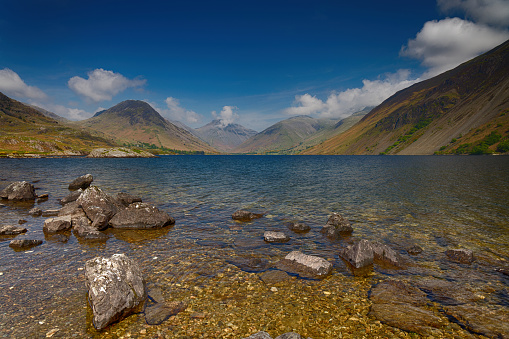 A view of Rocks at the foreshore of Wastwater in the Lake District Cumbria with Mountains in the background and white clouds on a Blue Sky