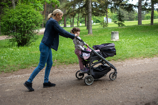 Grandma looks after her grandchildren. They walk in the forest, and push the baby stroller.