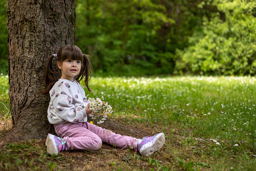 A little girl is playing in the forest with flowers. She made a beautiful bouquet, and now she rests on the ground.