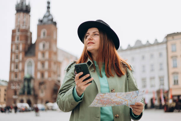 Beautiful stylish woman in hat walking on Market Square in Krakow on spring day and holding mobile phone and map. Phone Communication. Urban lifestyle concept. Check social networks, booking hotel hermosa mujer elegante con sombrero caminando por la plaza del mercado en cracovia el día de otoño y sosteniendo el teléfono móvil y el mapa. comunicación telefónica. concepto de estilo de vida urbano. consultar redes sociales, reserva hotel