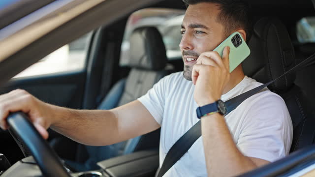Young hispanic man talking on smartphone sitting on car with winner expression at street