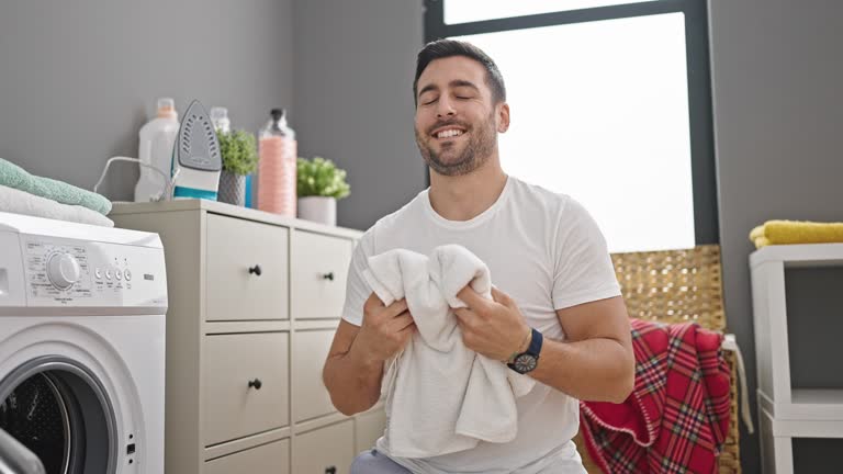 Young hispanic man smiling confident smelling clothes at laundry room