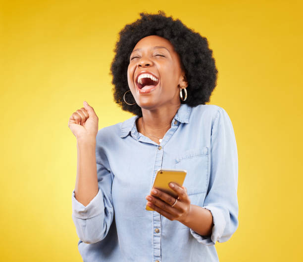 happy, excited and phone with black woman in studio for text message, notification and social media news. deal, winner and celebration with girl on yellow background for offer, giveaway and surprise - great discounts imagens e fotografias de stock