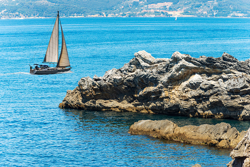 White sailing boat in the Mediterranean sea and rocky coast with cliffs in the Gulf of La Spezia, Liguria, Italy, Europe.