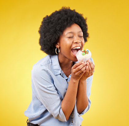 black woman, cupcake and excited or happy in studio while eating sweet food on a yellow background. African female model with snack, dessert or cake for happiness, birthday or celebration mockup