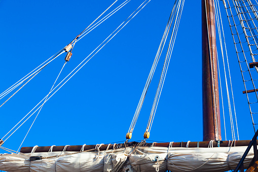 Part of an old, wooden sailing ship with a mast, folded sail and stretched sail ropes.