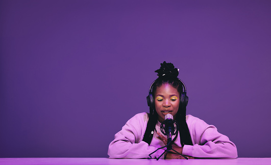 Female podcaster whispering into a microphone while hosting an asmr show. Woman recording a relaxing audio broadcast against a purple background. Black woman creating content for her internet podcast.