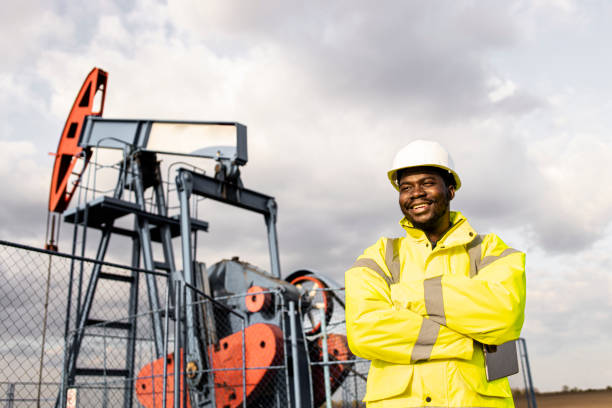 portret uśmiechniętego afroamerykańskiego robotnika w sprzęcie ochronnym stojącego przed platformami wiertniczymi. - oil industry digital tablet manual worker mining zdjęcia i obrazy z banku zdjęć