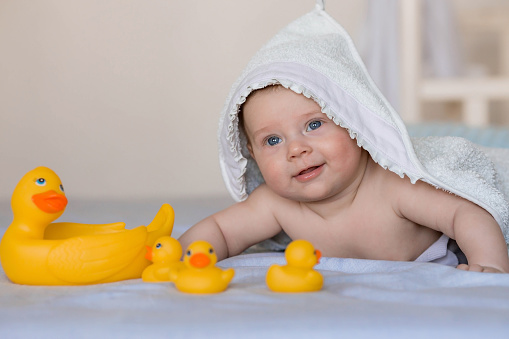 A charming newborn baby is lying on a bed with rubber ducks after bathing. A 4-month-old child with a towel on his head looks at the camera and smiles. Baby bathing and hygiene.