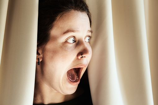 Peeking through window drapes, a young woman looks horrified with gaping mouth.