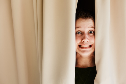 Peeking through window drapes, a young woman is terrified by what she sees.