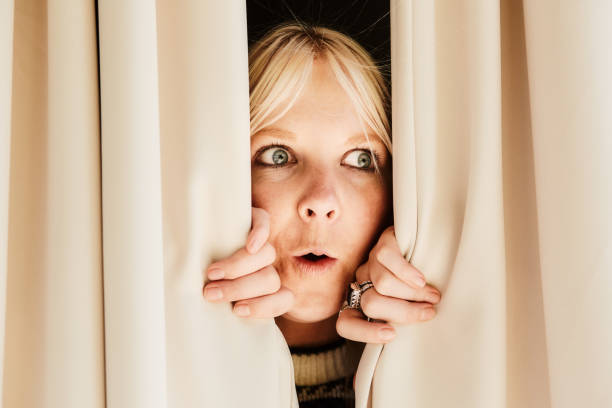 Young woman looking through curtains, her face a picture of surprise Peeking through window drapes, a young woman sees something she didn't expect. Hidden Meaning stock pictures, royalty-free photos & images