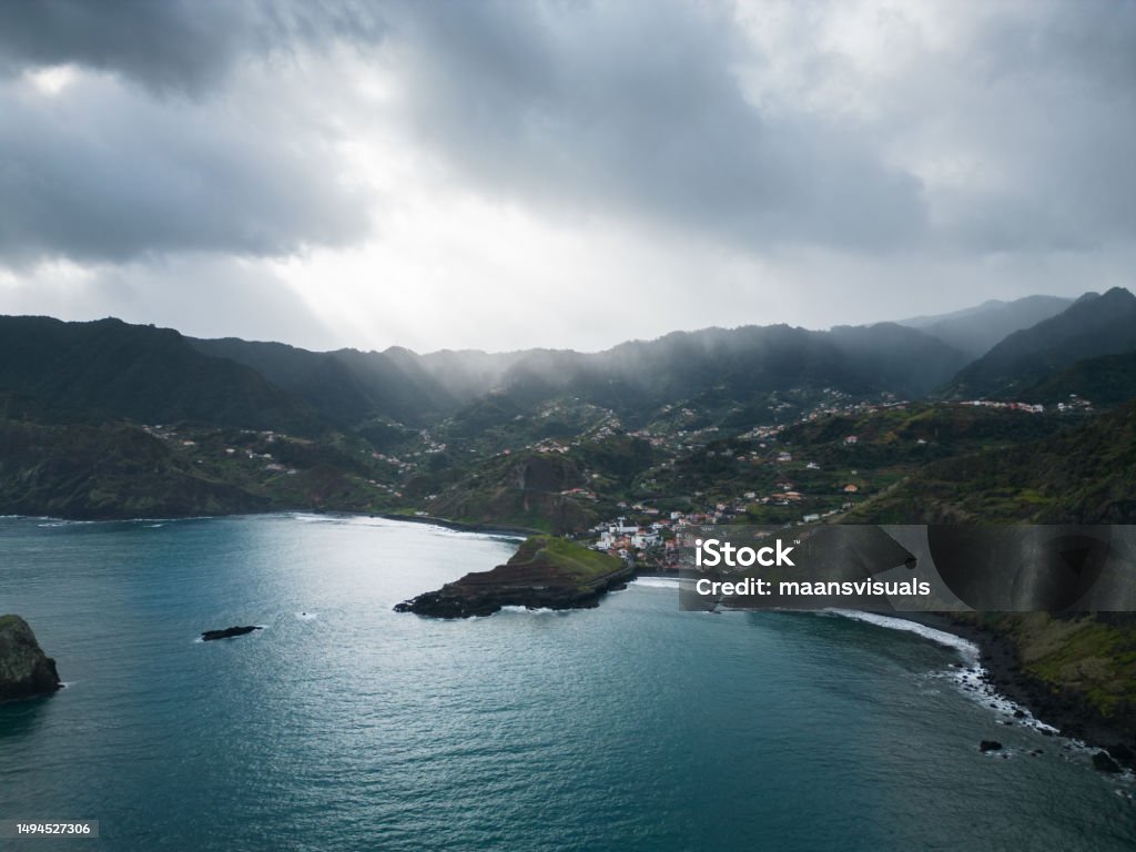 Madeira's hilly coastline village on the blue ocean in a cloudy day. Drone picture of the coastline of Madeira, Porto da Cruz, cloudy sky with sun rays peaking from the clouds, blue water and green land with villages and houses. Atlantic Islands Stock Photo