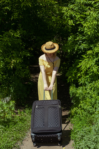Asian girl in straw hat and yellow dress with big black suitcase among green trees. Summer vacation. Time to travel