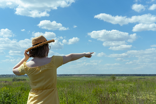 An Asian girl in a straw hat and a yellow dress points her finger at the sky with clouds in the field, rear view, copy space. Summer time. Village vacation. Time to travel