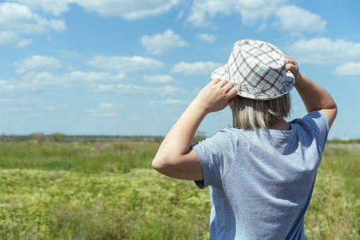 A woman in a hat and a blue T-shirt in a field, blue sky with clouds, rear view. Summer time. Village vacation. Time to travel