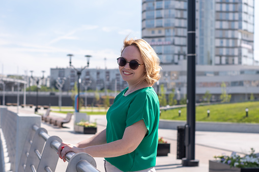 Portrait of a smiling happy girl in sunglasses, green blouse standing on the river bank. A beautiful confident student girl illuminated by the sun against the background of a blurred city.