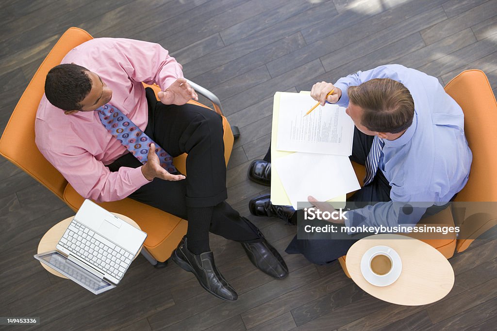 Due uomini d'affari seduti al chiuso con cartella e caffè portatile - Foto stock royalty-free di Riunione commerciale