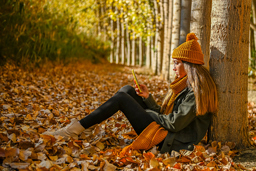Girl sitting among some autumn trees, looking at the mobile phone