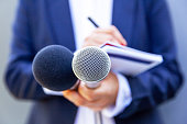 Microphone in the focus, news reporter at media event or press conference, holding mic, writing notes. Broadcast journalism concept.