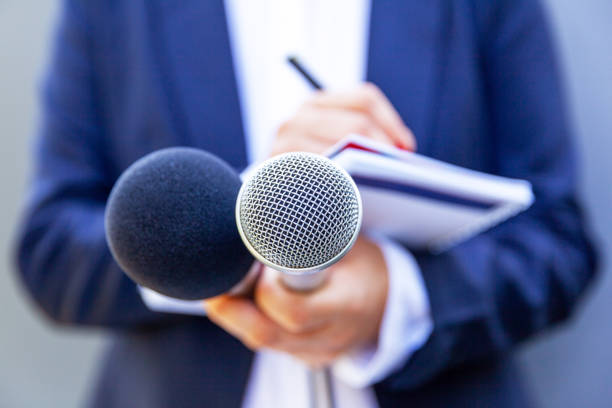 Microphone in the focus, news reporter at media event or press conference, holding mic, writing notes. Broadcast journalism concept. stock photo