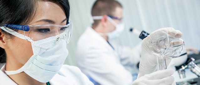 Chinese Asian woman female medical scientist, scientific researcher or doctor looking at a test tube and beaker of clear liquid in a medical research lab or laboratory with her colleague behind her. Panorama Web Banner Header