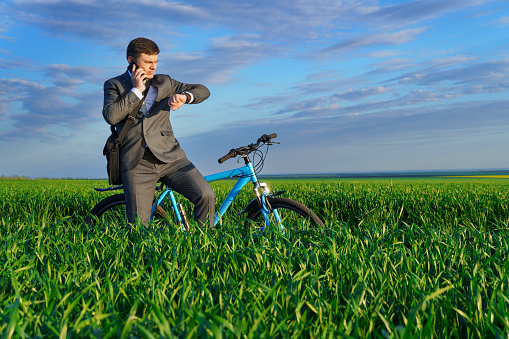 a businessman rides a bicycle on a green grassy field, dressed in a business suit, he makes a phone call and checks the time, beautiful nature in spring, freelance business concept