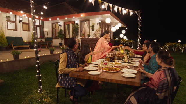 Big Family Celebrating Diwali: Indian Family in Traditional Clothes Gathered Together on a Dinner Table in a Backyard Garden Full of Lights. Moment of Happiness on a Hindu Holiday. Slow Motion