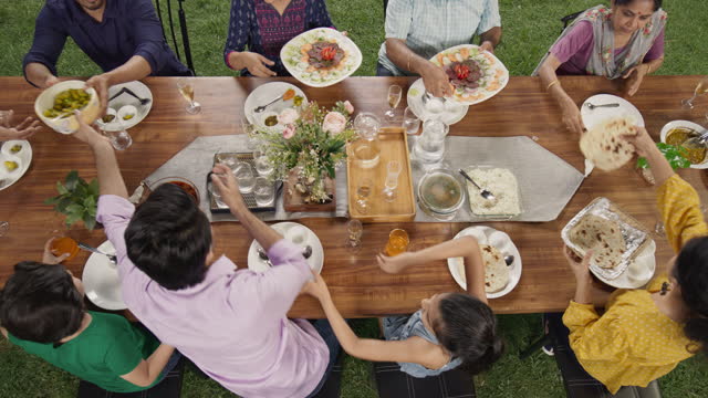 Big Indian Family Lunch Table: Top Down Elevated View at a Family and Friends Celebrating Outside at Home.Group of Children, Adults and Seniors Eating, Passing Traditional Dishes of Curry and Naan