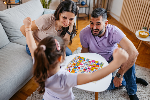 Young mother and father playing a Ludo board game with their daughter in the living room. Daughter winning in a game.
