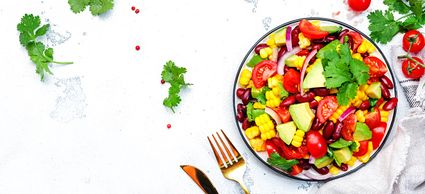 Spicy salad with sweet corn, red beans, avocado, jalapeno, cherry tomatoes, red onion and cilantro. White table background, top view banner