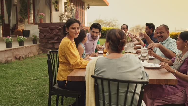 Indian Family Gathering Portrait: Family and Friends Celebrating Outside at Home. Diverse Group of Children, Adults and Seniors Sitting at a Table, Having Fun Conversations. Eating Traditional Food