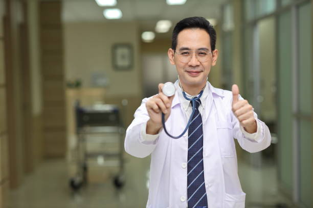 istock Happy asian medicine doctor standing and using stethoscope while showing thumb up and smile with hospital blurred background. Medical technology, treatment, healthcare and health insurance concept 1494550867