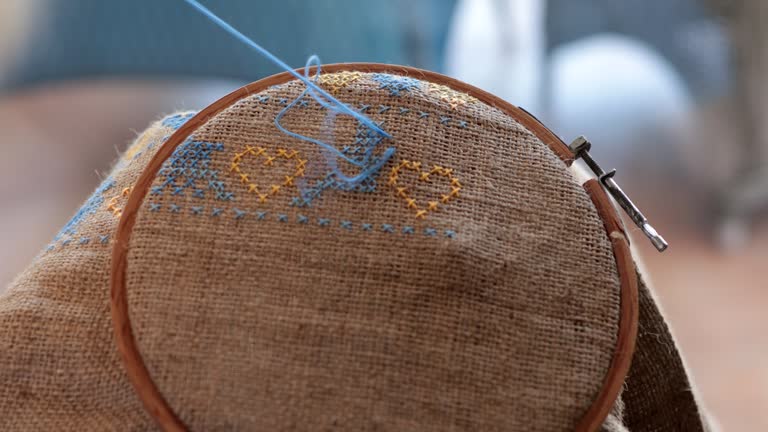 Fingers with needle embroider cross stitch blue bird on burlap with yellow hearts