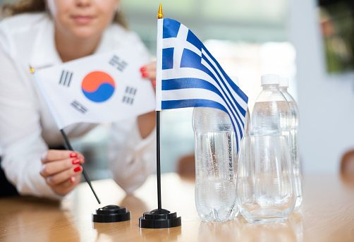 Young female assistant setting up greek and south korean flags for international negotiations