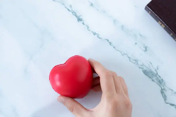 Human hand holding a red heart with closed holy bible book on white table. Top view, copy space. Love God Jesus Christ, Christian trust, obedience, and thanksgiving concept.