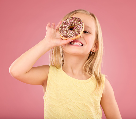 Child, donut and smile portrait in studio on a pink background while happy about sweet snack. Girl kid model with happiness, creativity and chocolate ring over eye in hand isolated on color and space