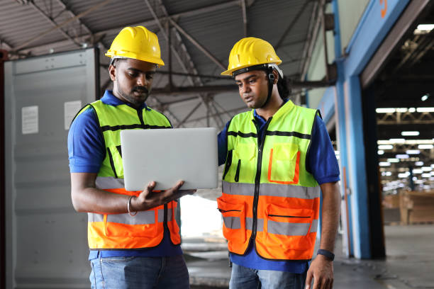 istock Group of warehouse workers with hardhats and reflective jackets using computer, walkie talkie radio and cardboard controlling stock and inventory in retail warehouse logistics, distribution center 1494568753